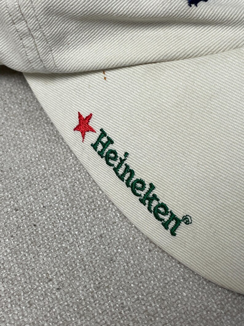 May include: A cream-colored baseball cap featuring an embroidered red star and the green text Heineken on the side of the brim. The fabric has a visible twill weave texture, and the stitching is consistent with standard cap construction.