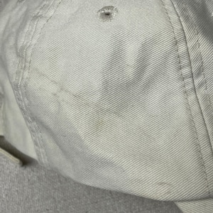 May include: A solid off-white baseball cap constructed from durable cotton twill fabric. The cap features a structured six-panel design with stitched eyelets on each panel for ventilation and reinforced seams running along the crown and brim.