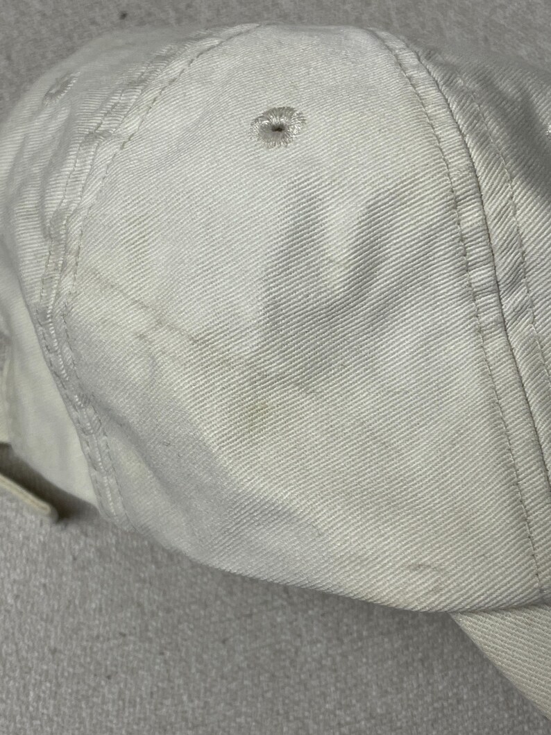 May include: A solid off-white baseball cap constructed from durable cotton twill fabric. The cap features a structured six-panel design with stitched eyelets on each panel for ventilation and reinforced seams running along the crown and brim.