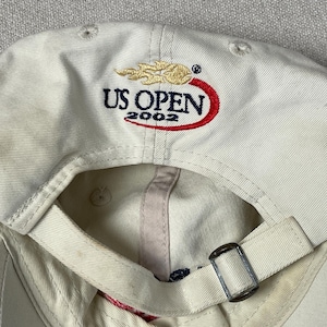 May include: An off-white baseball cap featuring an embroidered US OPEN 2002 logo on the back. The logo includes a gold flame graphic above the text and a red swoosh underneath. The cap has an adjustable fabric strap with a silver metal buckle.