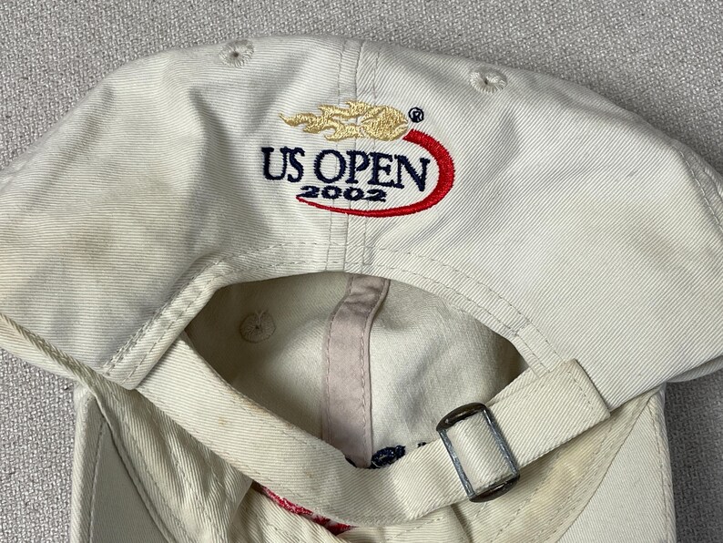 May include: An off-white baseball cap featuring an embroidered US OPEN 2002 logo on the back. The logo includes a gold flame graphic above the text and a red swoosh underneath. The cap has an adjustable fabric strap with a silver metal buckle.