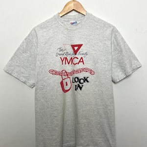 May include: A heather gray t-shirt with a graphic design featuring the text "The Grand River Area Family YMCA" in red and black. The design includes a red triangle, a chain, and the words "LOCK IN". The shirt is hanging on a wooden hanger.
