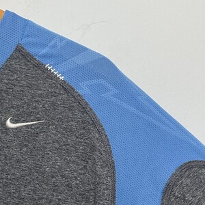 Puede incluir: Una camiseta deportiva gris y azul con el logo de Nike. La camiseta tiene un cuerpo gris con un panel de malla azul en el hombro y la manga. El logo de Nike es blanco.