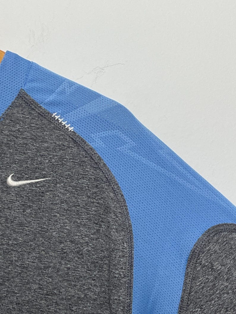 Puede incluir: Una camiseta deportiva gris y azul con el logo de Nike. La camiseta tiene un cuerpo gris con un panel de malla azul en el hombro y la manga. El logo de Nike es blanco.
