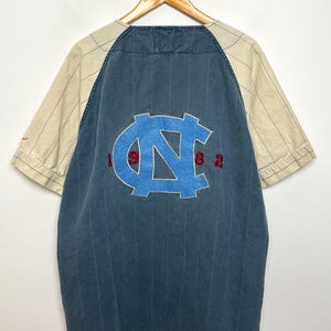 May include: A short-sleeve button-down shirt with a dark blue pinstriped main body and cream-colored pinstriped sleeves. The back features a large light blue embroidered NC logo with the red embroidered numbers 1, 9, 8, and 2 flanking it.
