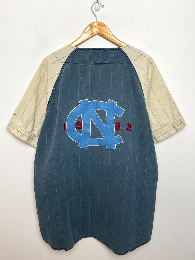 May include: A short-sleeve button-down shirt with a dark blue pinstriped main body and cream-colored pinstriped sleeves. The back features a large light blue embroidered NC logo with the red embroidered numbers 1, 9, 8, and 2 flanking it.