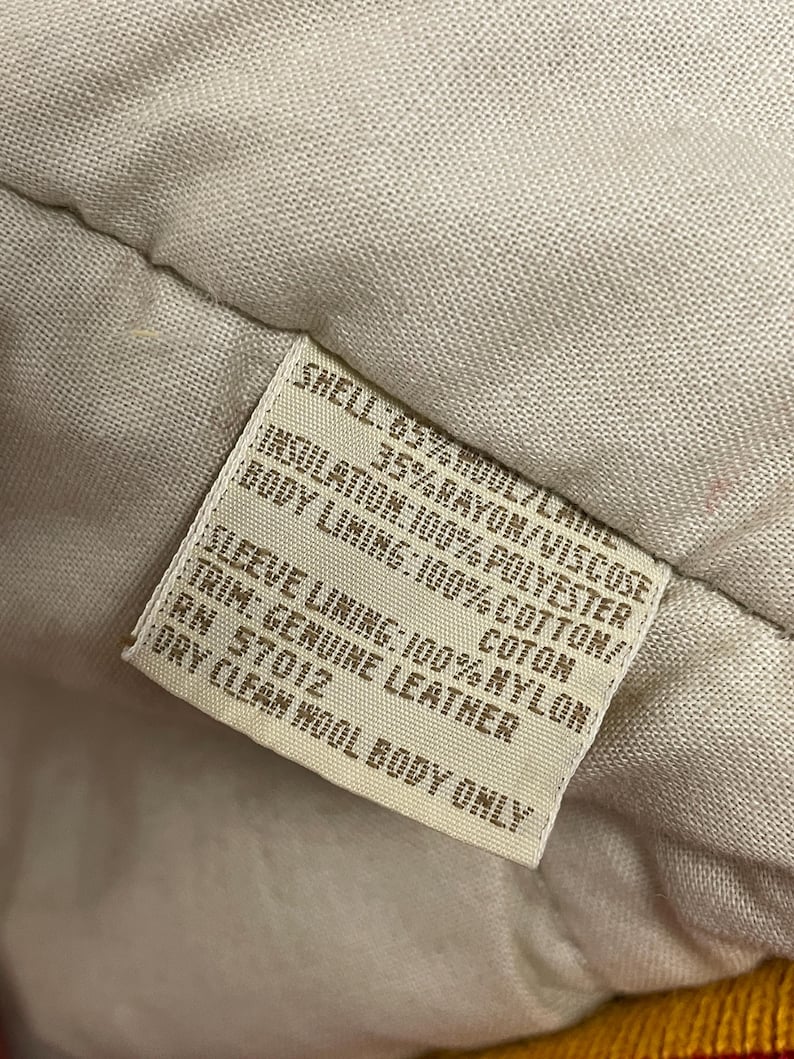 May include: A close-up of a clothing label detailing the materials used. The shell is 65% wool and 35% rayon/viscose. The insulation is polyester, the body lining is cotton, and the sleeve lining is nylon. The trim is genuine leather.