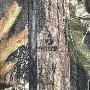 Pode incluir: Close-up de um tecido camuflado com um z&iacute;per preto no centro. O tecido apresenta uma mistura de cores marrom, verde e bege, com o texto "Mossy Oak Break Up" vis&iacute;vel.