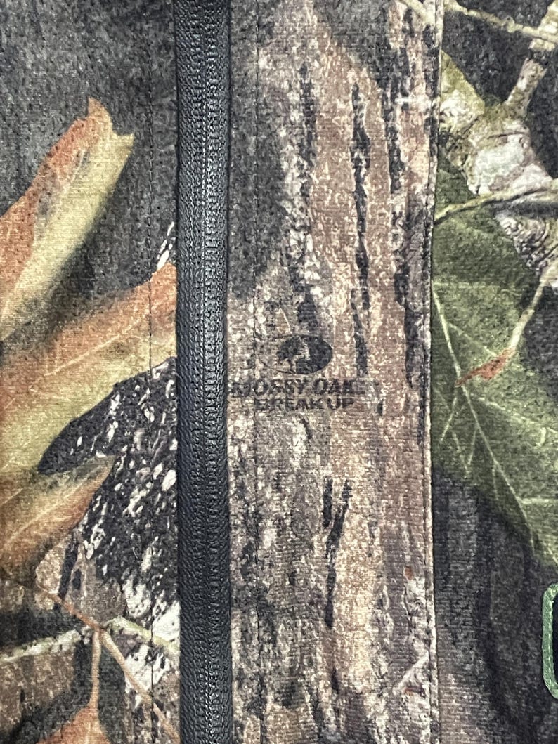 Pode incluir: Close-up de um tecido camuflado com um z&iacute;per preto no centro. O tecido apresenta uma mistura de cores marrom, verde e bege, com o texto "Mossy Oak Break Up" vis&iacute;vel.