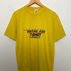 May include: A yellow T-shirt with the text "BLAZE SSI T-SHIRT VERSION 1.0" in black, red, and gold lettering. The shirt is hanging on a wooden hanger against a white background. The shirt has short sleeves and a crew neck.