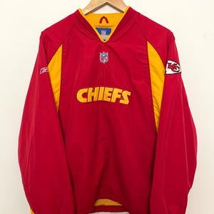 Puede incluir: Chaqueta tipo jersey de los Chiefs en rojo y amarillo. La chaqueta presenta el logotipo de los Chiefs en el pecho y el logotipo del equipo en la manga. La chaqueta tiene cuello en V y est&aacute; colgada en una percha de madera.