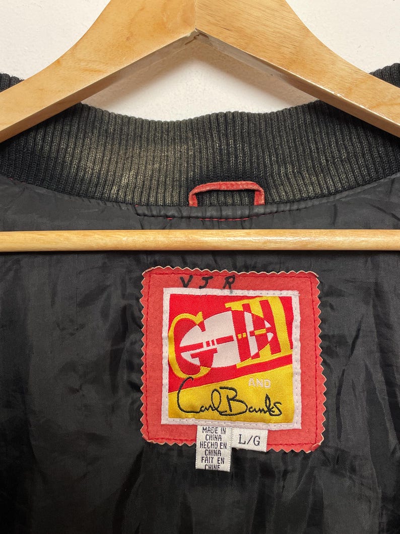 Puede incluir: Una chaqueta de cuero roja y negra con una etiqueta roja que dice "Carl Banks" y "VIR" en letras blancas. La etiqueta tambi&eacute;n dice "Made in China, Hecho en China, Fait en Chine" y "L/G".