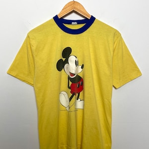 K&ouml;nnte beinhalten: Gelbes T-Shirt mit blauem Kragen und B&uuml;ndchen, mit einer Mickey Mouse-Grafik. Die Zeichentrickfigur ist in Schwarz, Rot, Wei&szlig; und Beige dargestellt. Das T-Shirt h&auml;ngt an einem Holzkleiderb&uuml;gel.