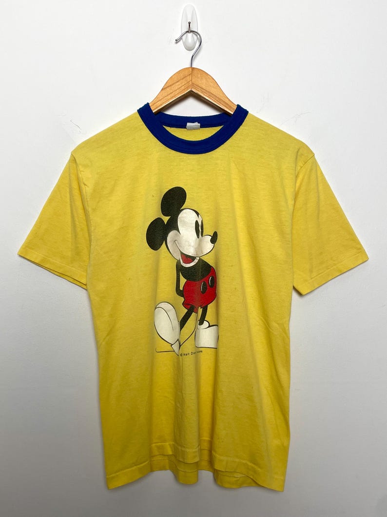 K&ouml;nnte beinhalten: Gelbes T-Shirt mit blauem Kragen und B&uuml;ndchen, mit einer Mickey Mouse-Grafik. Die Zeichentrickfigur ist in Schwarz, Rot, Wei&szlig; und Beige dargestellt. Das T-Shirt h&auml;ngt an einem Holzkleiderb&uuml;gel.
