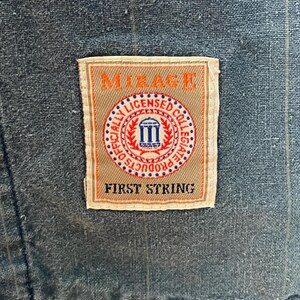 May include: A blue fabric garment with a subtle pinstripe pattern featuring a rectangular patch label. The label displays the text MIRAGE at the top, FIRST STRING at the bottom, and a central circular emblem reading OFFICIALLY LICENSED COLLEGIATE PRODUCTS.