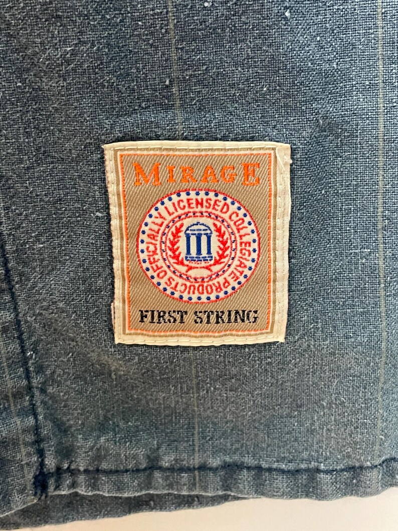 May include: A blue fabric garment with a subtle pinstripe pattern featuring a rectangular patch label. The label displays the text MIRAGE at the top, FIRST STRING at the bottom, and a central circular emblem reading OFFICIALLY LICENSED COLLEGIATE PRODUCTS.
