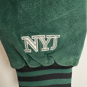 Pode incluir: Close-up de uma manga de casaco de camur&ccedil;a verde. A manga apresenta as letras bordadas "NYJ" em branco. O punho &eacute; listrado em preto e verde.