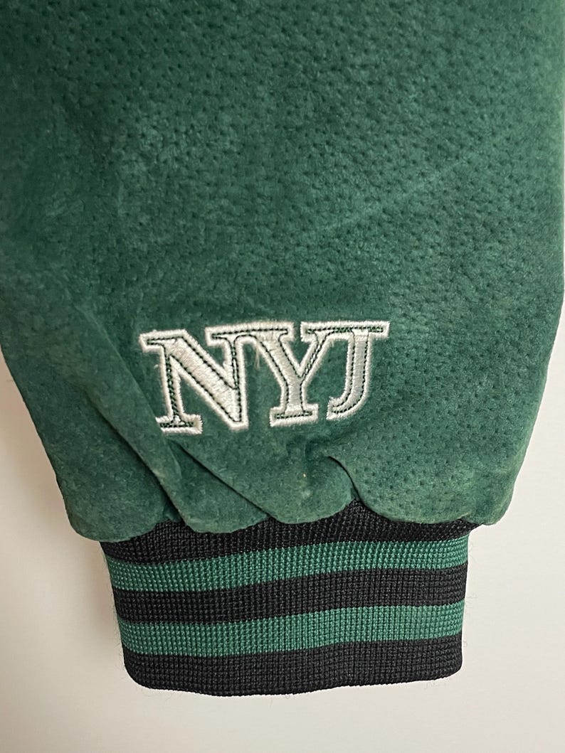 Pode incluir: Close-up de uma manga de casaco de camur&ccedil;a verde. A manga apresenta as letras bordadas "NYJ" em branco. O punho &eacute; listrado em preto e verde.