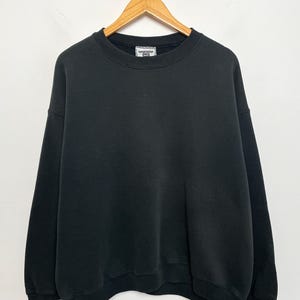 May include: A black crew neck sweatshirt hanging on a wooden hanger. The sweatshirt has a ribbed collar, cuffs, and hem. The garment is made of a thick, soft material. The background is a plain white wall.