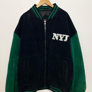 Pode incluir: Casaco universit&aacute;rio preto com mangas e acabamentos verdes. O casaco apresenta as letras "NYJ" em branco no peito. O casaco tem fecho de correr e est&aacute; pendurado num cabide.