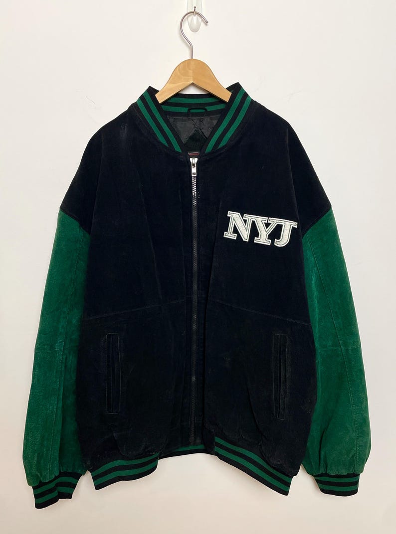 Pode incluir: Casaco universit&aacute;rio preto com mangas e acabamentos verdes. O casaco apresenta as letras "NYJ" em branco no peito. O casaco tem fecho de correr e est&aacute; pendurado num cabide.