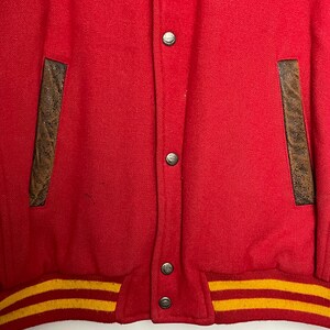 May include: A red varsity jacket with brown leather pocket details and a yellow and gold striped hem. The jacket features silver snap closures.