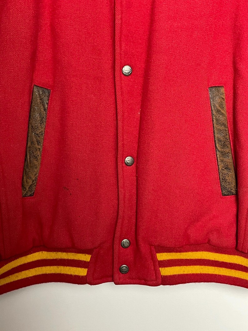 May include: A red varsity jacket with brown leather pocket details and a yellow and gold striped hem. The jacket features silver snap closures.