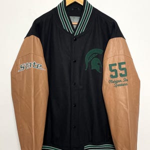 Puede incluir: Chaqueta universitaria negra y marrón con detalles de rayas verdes y blancas. La manga izquierda muestra "State", la manga derecha tiene "55" y "Michigan State Spartans". Un logotipo de casco espartano verde está en el pecho.
