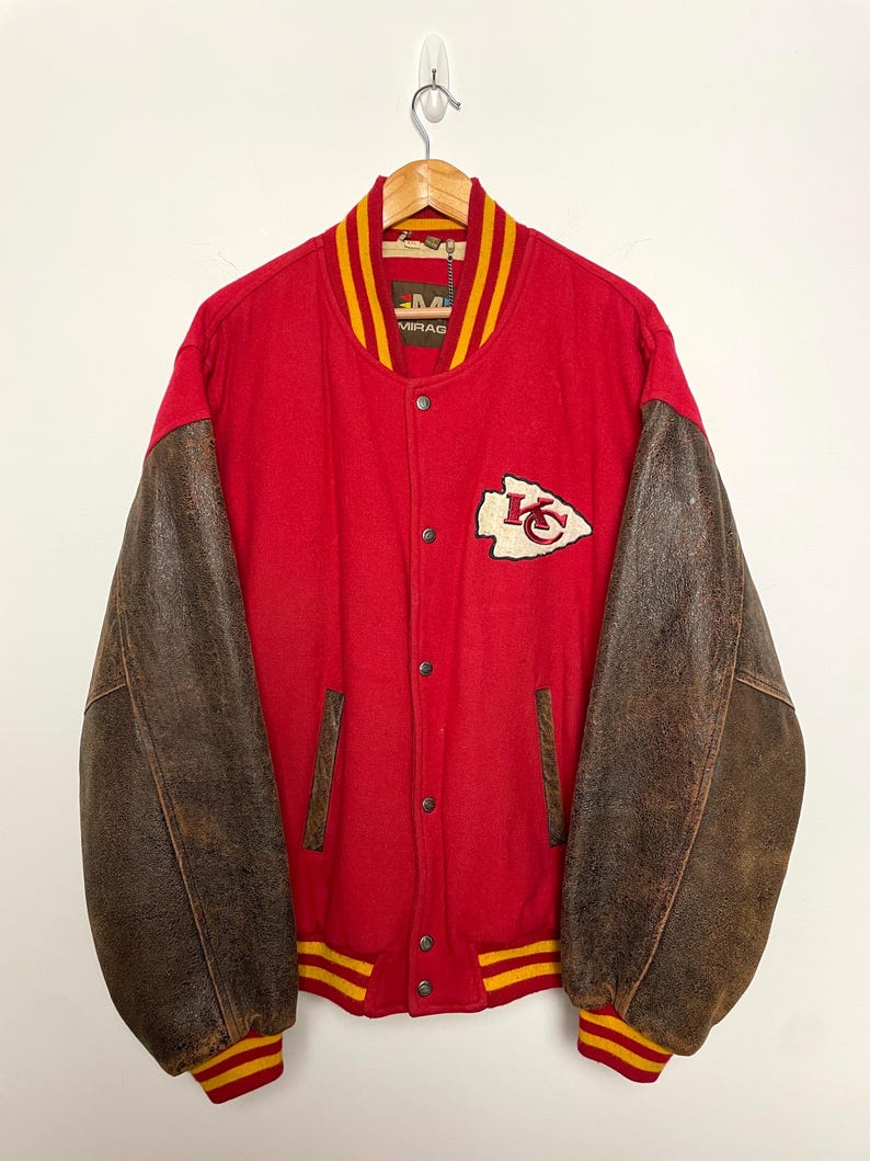 May include: A vintage letterman jacket in red and brown, with a snap-button closure. It has brown leather sleeves and a red body with yellow and red striped trim. A team logo patch is on the chest.