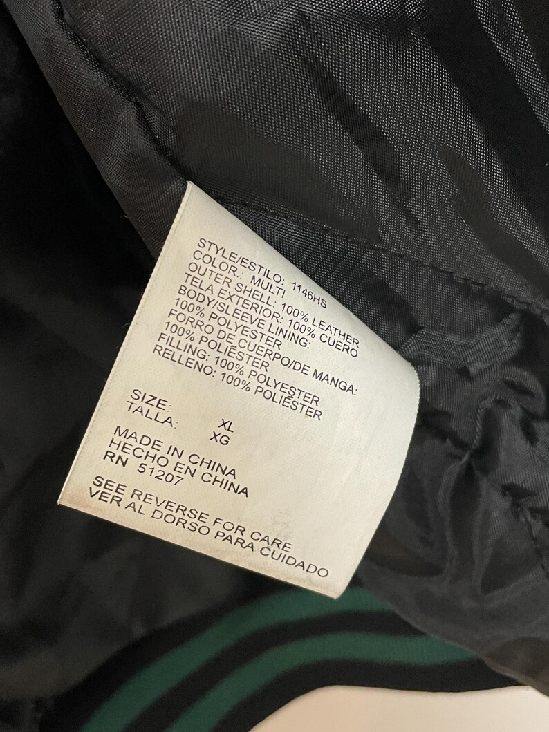 Pode incluir: Close-up de uma etiqueta de jaqueta de couro preta com texto. A etiqueta indica que a jaqueta &eacute; multicolorida, feita de 100% couro e tamanho XL. A etiqueta tamb&eacute;m afirma que a jaqueta foi feita na China.