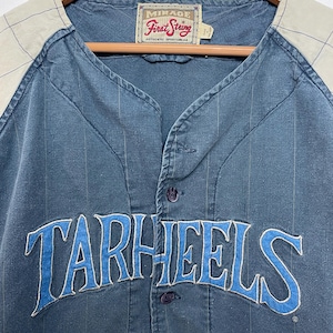 May include: A blue button-down baseball jersey with thin vertical pinstripes and light beige raglan sleeves featuring a subtle grid pattern. The word TARHEELS is embroidered in blue with white trim across the chest. A Mirage First String label is at the neck.