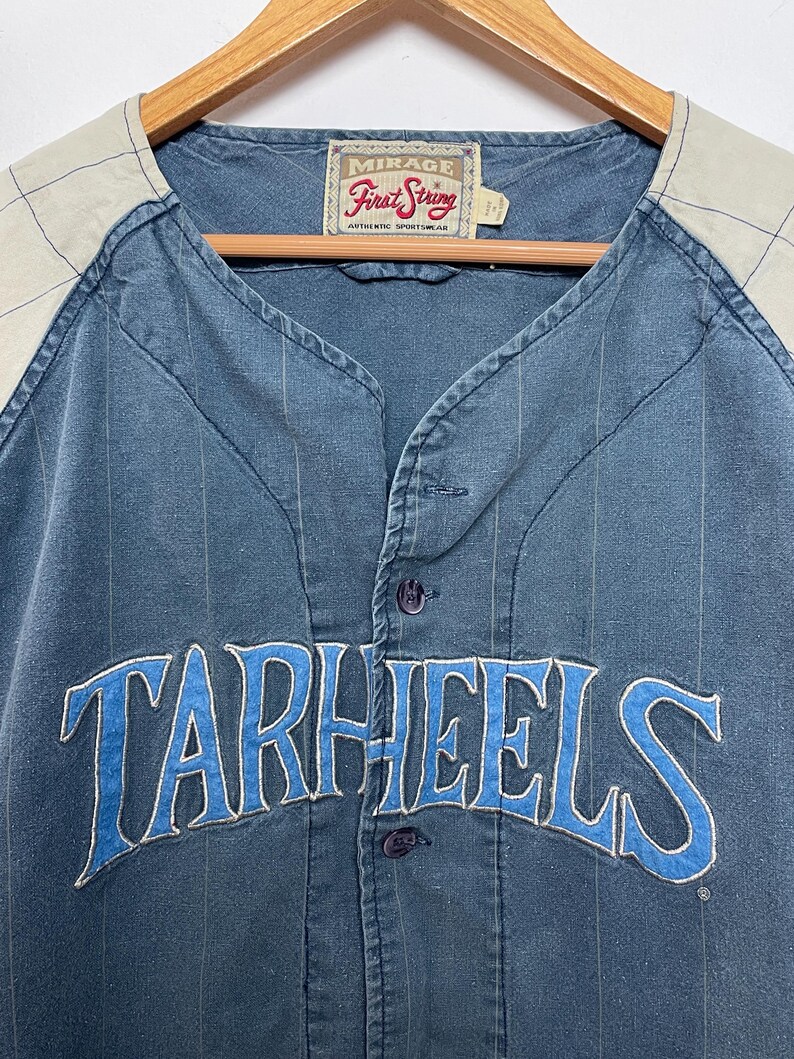 May include: A blue button-down baseball jersey with thin vertical pinstripes and light beige raglan sleeves featuring a subtle grid pattern. The word TARHEELS is embroidered in blue with white trim across the chest. A Mirage First String label is at the neck.