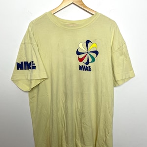 May include: Pale yellow t-shirt featuring the Nike logo on the left sleeve and chest. The chest logo is a colourful flower design with the word "NIKE" below it. The sleeve has the word "NIKE" in blue.