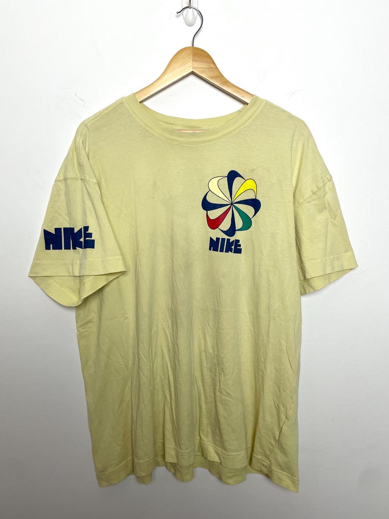 May include: Pale yellow t-shirt featuring the Nike logo on the left sleeve and chest. The chest logo is a colourful flower design with the word "NIKE" below it. The sleeve has the word "NIKE" in blue.