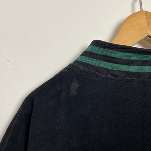 Pode incluir: Close-up de uma jaqueta preta com superf&iacute;cie texturizada. A gola apresenta listras verdes e pretas. A jaqueta est&aacute; pendurada em um cabide de madeira contra uma parede branca.