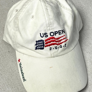 May include: An off-white baseball cap featuring embroidered text that reads US OPEN and 2.0.0.2, accompanied by a stylized American flag graphic. The brim displays a red star and the green text Heineken embroidered on the left side.