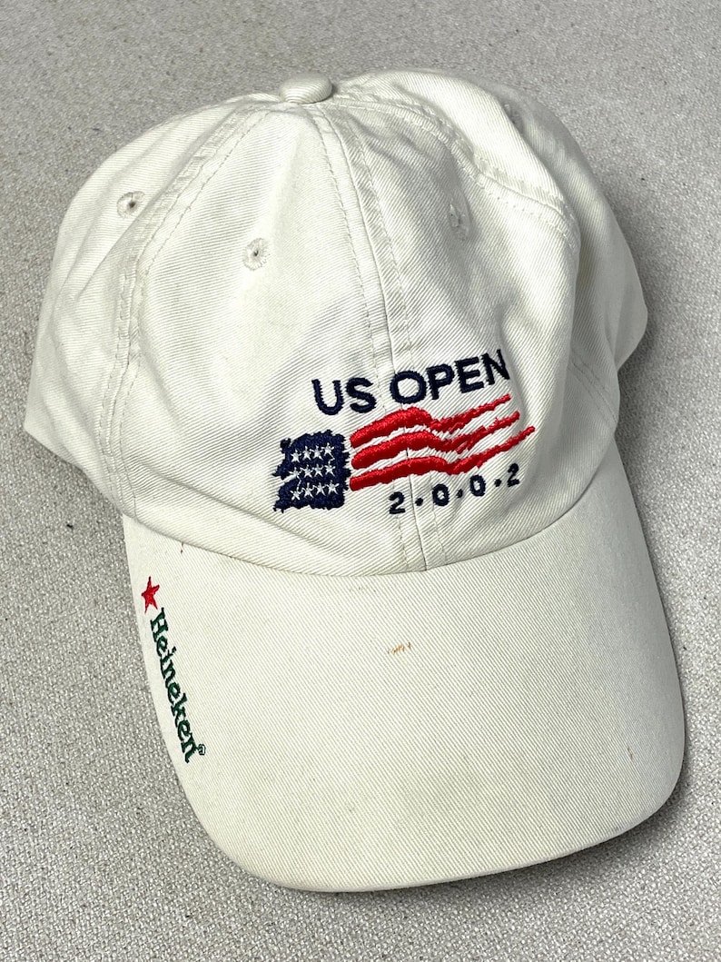 May include: An off-white baseball cap featuring embroidered text that reads US OPEN and 2.0.0.2, accompanied by a stylized American flag graphic. The brim displays a red star and the green text Heineken embroidered on the left side.