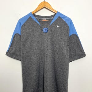 Puede incluir: Camiseta gris de manga corta con paneles en los hombros de color azul claro y un logotipo azul claro en el pecho. El logotipo de Nike es visible en el pecho derecho. La camiseta est&aacute; colgada en una percha de madera.