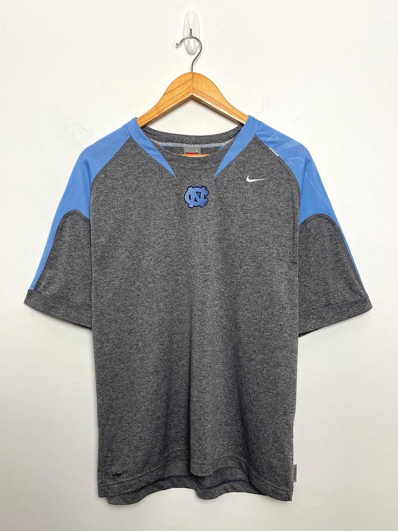 Puede incluir: Camiseta gris de manga corta con paneles en los hombros de color azul claro y un logotipo azul claro en el pecho. El logotipo de Nike es visible en el pecho derecho. La camiseta est&aacute; colgada en una percha de madera.