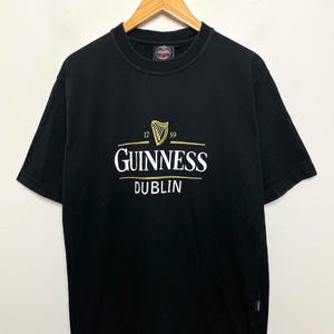 May include: Black t-shirt featuring the Guinness logo in gold and white, with a harp emblem and the words "GUINNESS DUBLIN". The shirt is displayed on a wooden hanger.