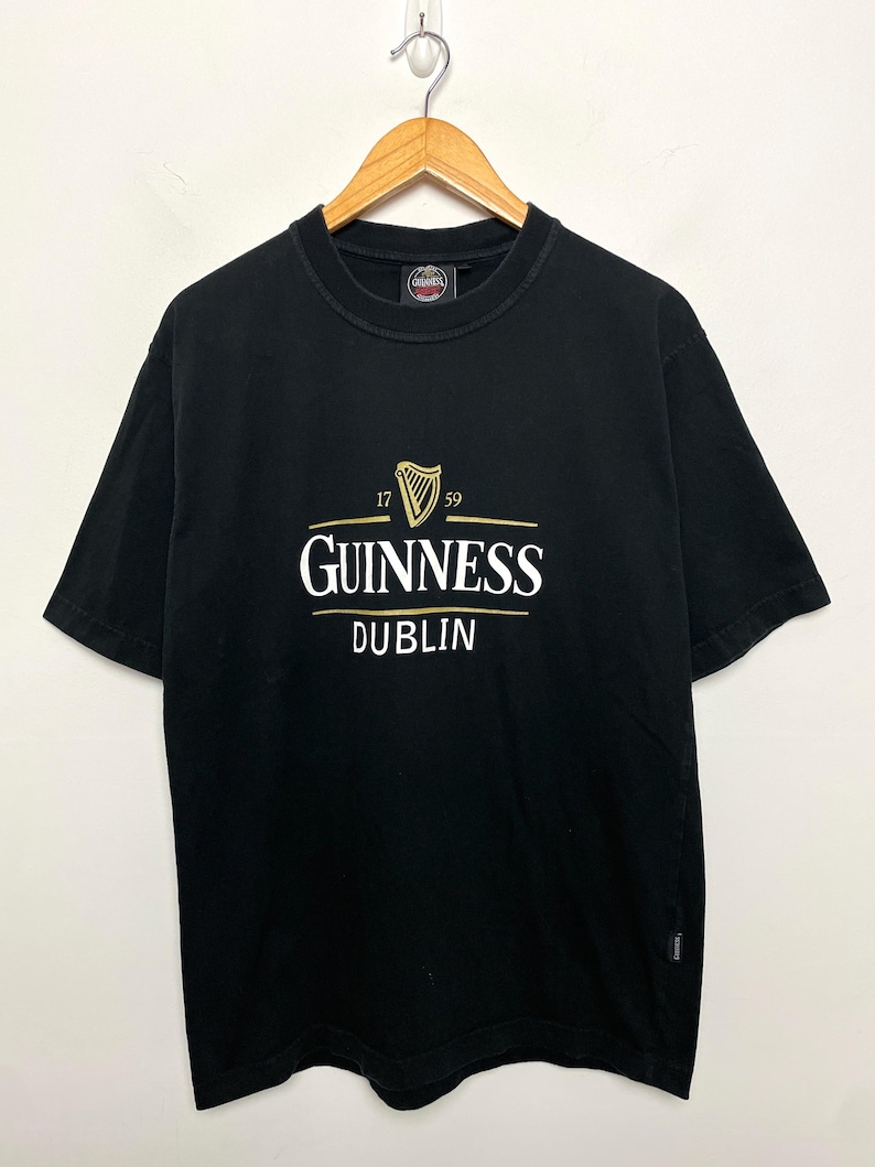 May include: Black t-shirt featuring the Guinness logo in gold and white, with a harp emblem and the words "GUINNESS DUBLIN". The shirt is displayed on a wooden hanger.