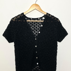 May include: A black, short-sleeved cardigan with a textured, open-weave design. The button-down top features a V-neck and is displayed on a wooden hanger against a white background. The cardigan has small, round, white buttons.