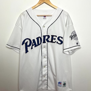 Può includere: Maglia da baseball bianca con la scritta "PADRES" in blu navy sul petto. La maglia ha finiture blu navy e una toppa con il logo della squadra sulla manica. La maglia è abbottonata e ha un'etichetta Russell Athletic.