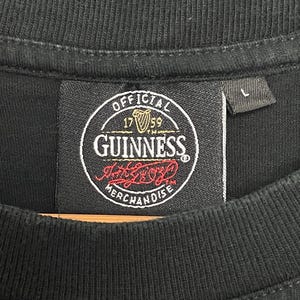 May include: Black t-shirt featuring a Guinness label. The label is circular and displays the Guinness logo, the words "OFFICIAL MERCHANDISE", and the year "1759". A size "L" tag is visible.