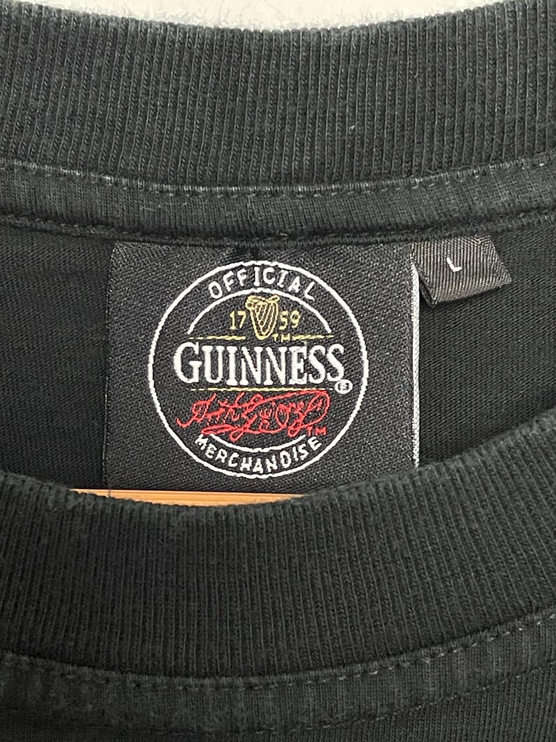 May include: Black t-shirt featuring a Guinness label. The label is circular and displays the Guinness logo, the words "OFFICIAL MERCHANDISE", and the year "1759". A size "L" tag is visible.