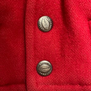 May include: Close-up of red fabric with two silver buttons. Each button is round and features the words "National Football League" and a football shape. The fabric has a textured appearance.