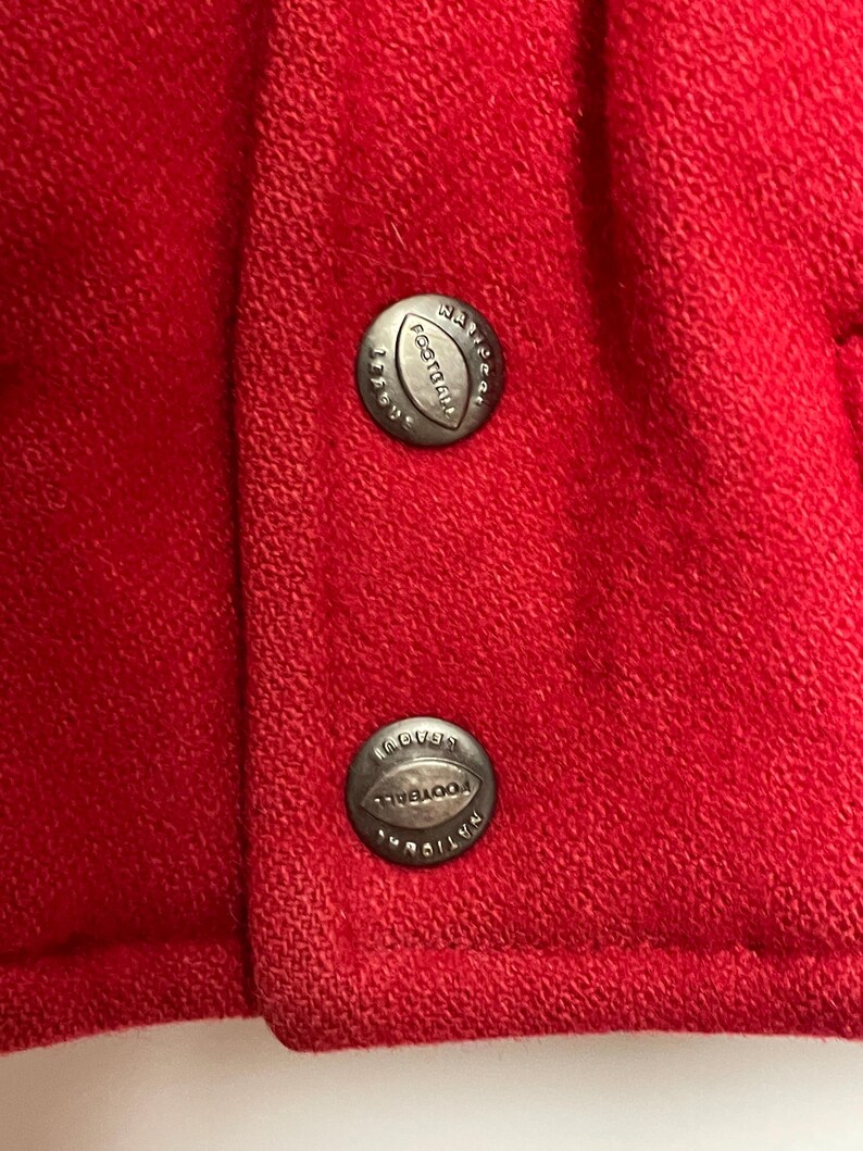 May include: Close-up of red fabric with two silver buttons. Each button is round and features the words "National Football League" and a football shape. The fabric has a textured appearance.