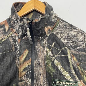 Pode incluir: Casaco de ca&ccedil;a camuflado com z&iacute;per preto de comprimento total e gola alta. O casaco apresenta um patch verde e branco "MEDALIST HUNTGEAR". O padr&atilde;o inclui tons de castanho, verde e cinzento.