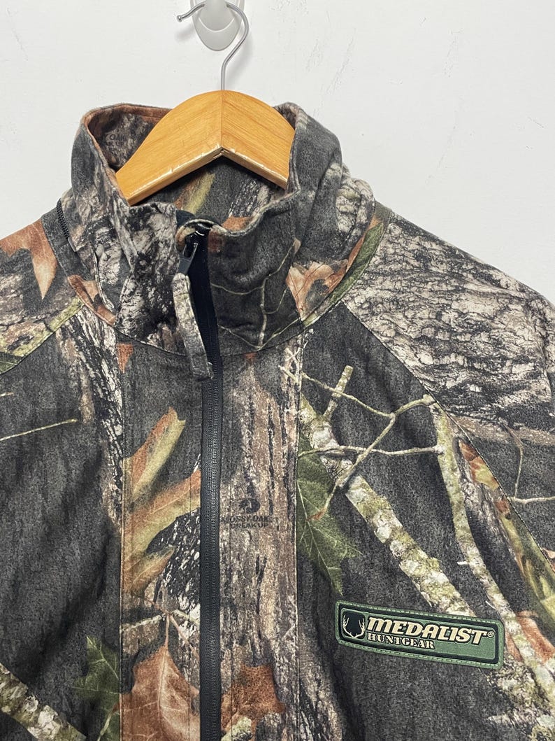 Pode incluir: Casaco de ca&ccedil;a camuflado com z&iacute;per preto de comprimento total e gola alta. O casaco apresenta um patch verde e branco "MEDALIST HUNTGEAR". O padr&atilde;o inclui tons de castanho, verde e cinzento.