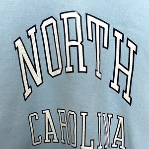 K&ouml;nnte beinhalten: Hellblauer Pullover mit wei&szlig;er und schwarzer Schrift, die "NORTH CAROLINA" lautet.